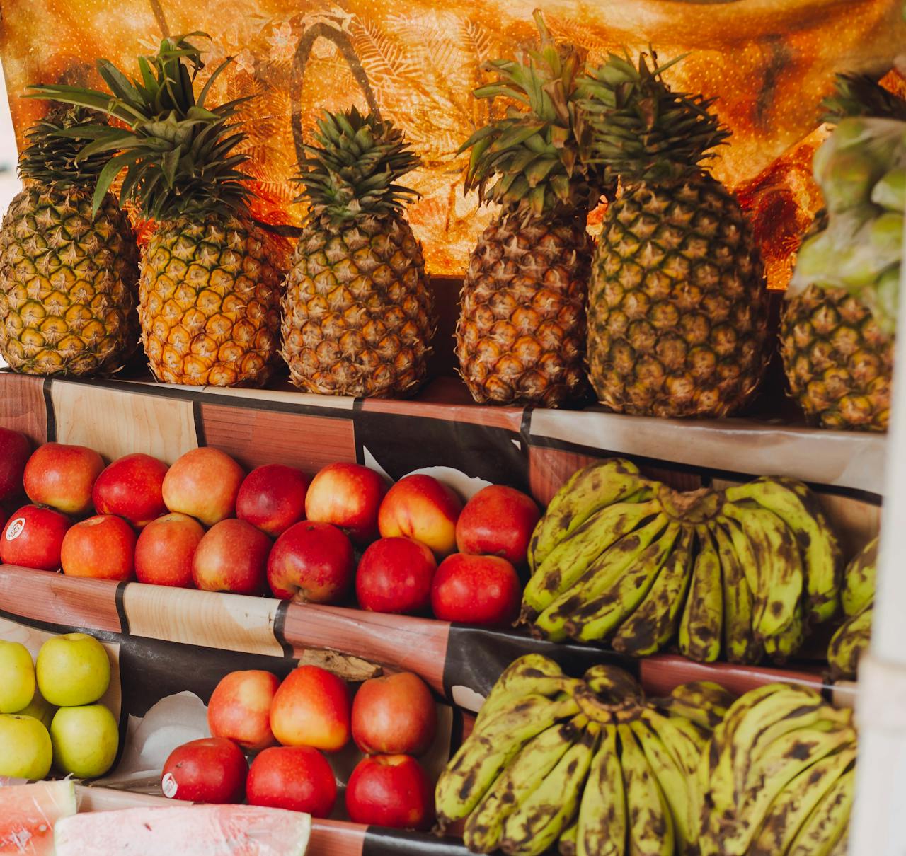 services-02 A colorful display of pineapples, apples, and plantains at a fruit market stand.